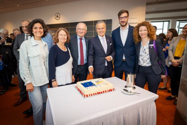 Referenten stehen vor einer cremefarbenen Torte mit blauem, gelbem und rotem Zuckerdecor